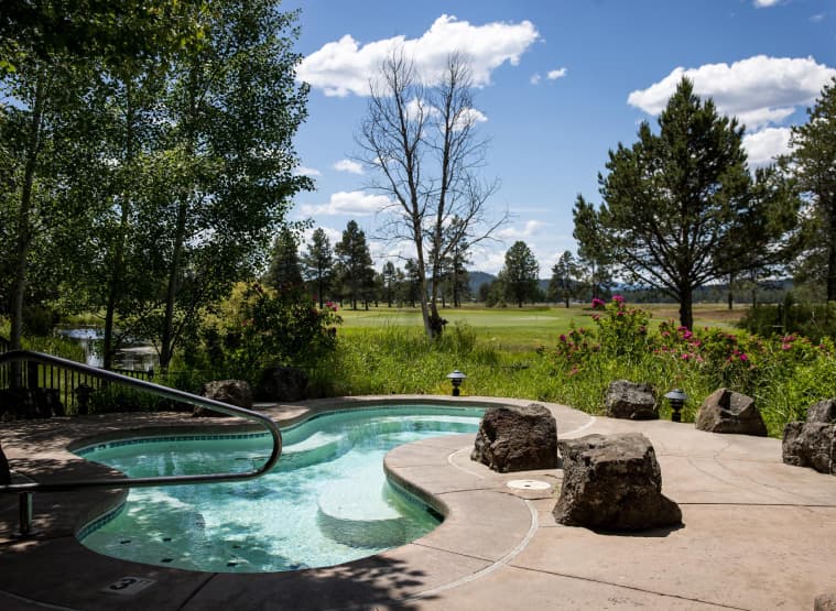 A small outdoor hot tub surrounded by rocks and trees overlooks a grassy field and golf course under a blue sky with scattered clouds.