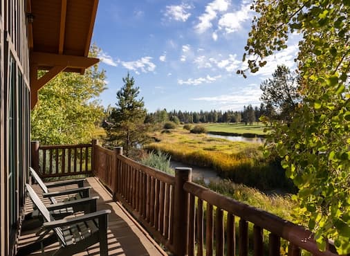 A wooden deck with chairs overlooks a scenic meadow, pond, and trees under a partly cloudy blue sky, suggesting a peaceful and natural setting.
