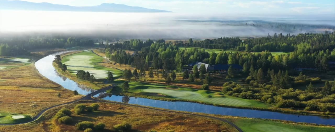 A golf course with green fairways, a winding river, trees, and a bridge, surrounded by forest and mountains in the distance under a cloudy sky with mist.