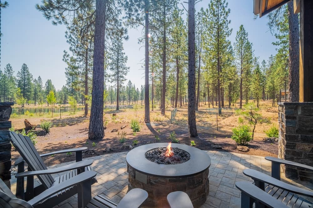 Four Adirondack chairs surround a round fire pit on a stone patio, overlooking a pine forest under a clear sky.