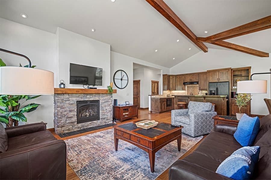 A cozy living room with a stone fireplace, TV, brown leather sofas, wood beams, and an open kitchen with dark cabinets in the background.