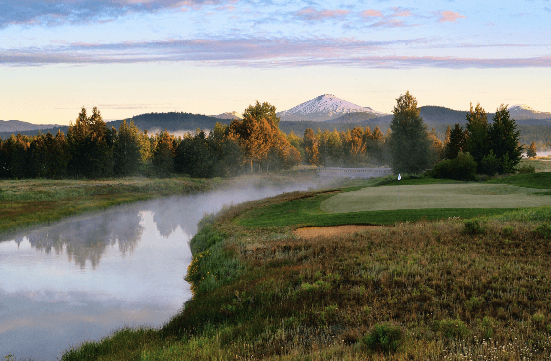 A golf green with a flag sits beside a misty river, surrounded by trees and grass, with snow-capped mountains and a colorful sky in the background.