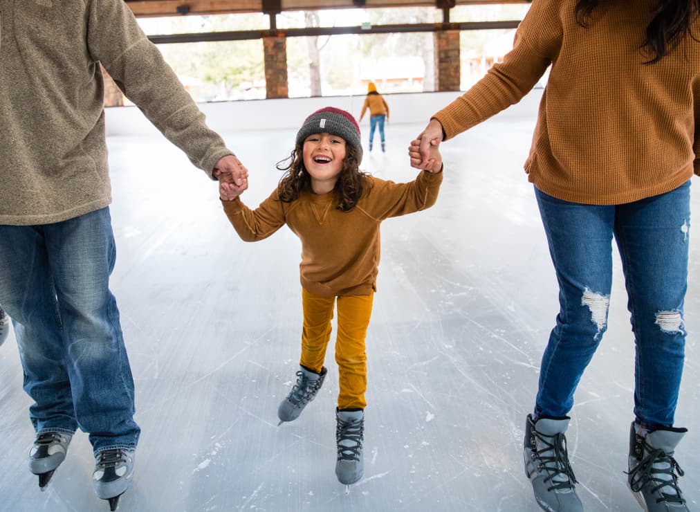 A smiling child in a gray beanie and brown sweater ice skates while holding hands with two adults, all wearing jeans or casual pants, on an indoor ice rink.