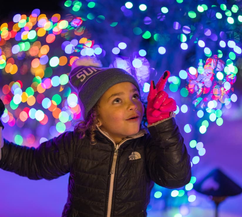 A young child in a winter jacket and hat points excitedly at colorful holiday lights, with a joyful expression and bright bokeh in the background.