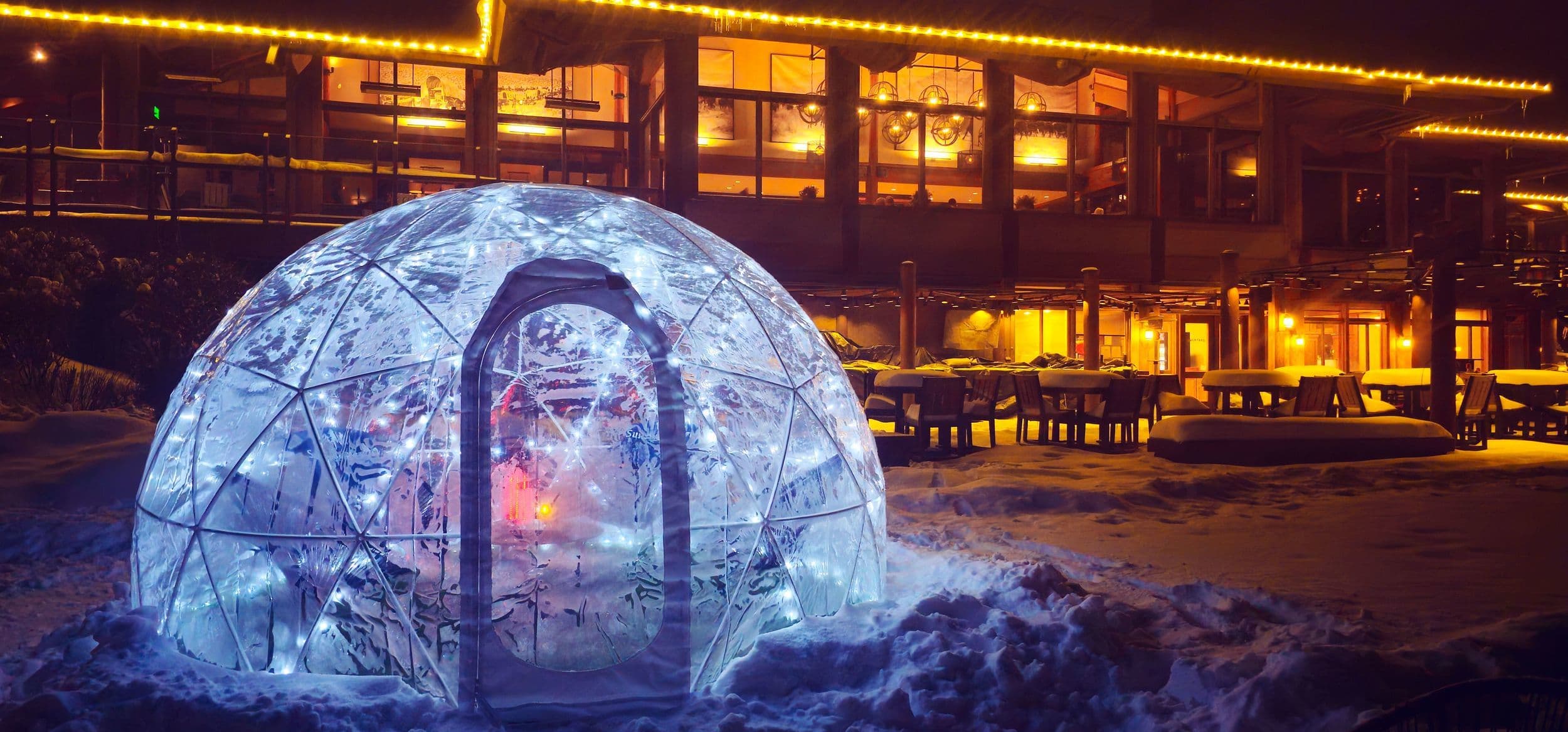 A glowing transparent igloo sits in the snow outside a warmly lit building with string lights, tables, and chairs visible on a winter night.