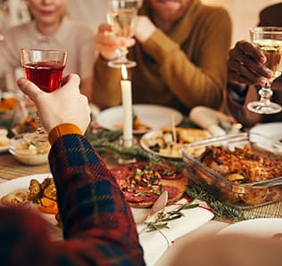 Four people sit around a festive dinner table, raising glasses in a toast. The table is decorated with greenery and candles, and is set with various dishes, including a casserole and roasted vegetables.