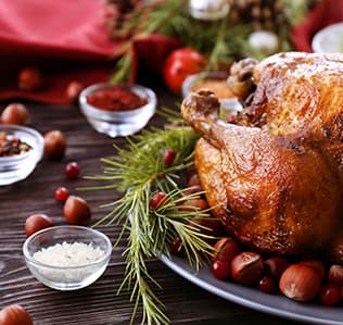 Roast chicken on a plate garnished with pine branches, cranberries, and hazelnuts, surrounded by small bowls of spices and salt on a wooden table with festive decor in the background.