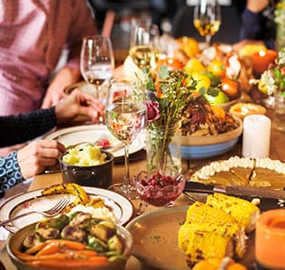 A group of people enjoy a festive meal with wine, mashed potatoes, corn, pie, vegetables, and flowers on a wooden table, creating a warm, inviting atmosphere.