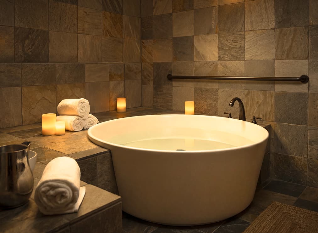 Private spa soaking tub surrounded by stone tile, rolled towels, and softly glowing candles.